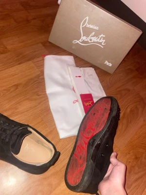 Svarta Louboutin sneakers med nitar - Säljer ett par svarta sneakers från Christian Louboutin med klassisk röd sula och svarta nitar på tån. Skorna är i mocka och har svarta skosnören. Kommer med originalkartong och dustbag. Perfekta för dig som vill sticka ut med en lyxig och edgy stil.