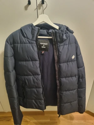 Jacka  - Snygg mörkblå pufferjacka från Superdry med huva och dragkedja framtill. Jackan har två fickor med dragkedja och ett diskret logomärke på bröstet. 