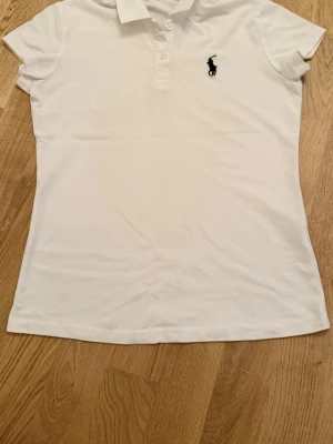 Vit pikétröja från Ralph Lauren - Klassisk vit pikétröja från Ralph Lauren med korta ärmar och krage. Tröjan har knäppning framtill och den ikoniska broderade loggan på bröstet. Perfekt för en clean och sportig look.
