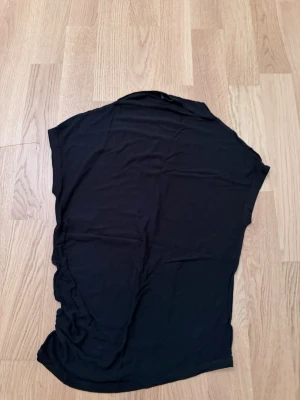 Svart topp med rynkade detaljer - Svart topp i mjukt material med rynkade detaljer längs ena sidan. Modellen är ärmlös och har en enkel, stilren design som passar till många olika looks. Perfekt för dig som gillar minimalistisk stil med en twist.