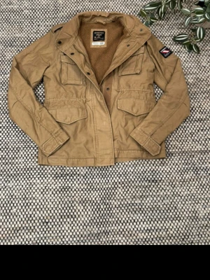 Beige fieldjacka med fickor från Superdry - Stilren beige field från Superdry med fyra stora fickor framtill och patch på ärmen. Jackan har en mysig teddyfodrad insida och klassisk krage. Perfekt för dig som gillar en avslappnad och trendig look.