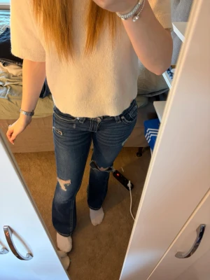 Blå bootcut jeans med slitningar - Snygga blå bootcut jeans med slitningar på knä och lår. Jeansen har låg midja, klassiska fem fickor och kontrastsömmar. Perfekta för en avslappnad och trendig look. Storlek Gina Y 14+ S tall. Passar mig som är 175cm