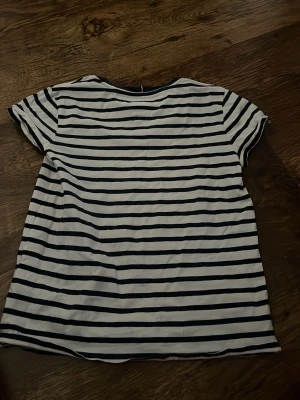 Randig T-shirt från Tommy Hilfiger - Vit och marinblå randig T-shirt från Tommy Hilfiger med korta ärmar och rund hals. T-shirten har logotypen broderad framtill och är gjord i mjuk bomull. Perfekt för en avslappnad och stilren look.