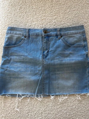 Blå jeanskjol med fransig kant - Snygg blå jeanskjol i klassisk modell med fem fickor, bälteshällor och rå, fransig nederkant. Kjolen har knapp och dragkedja framtill och är perfekt för en avslappnad stil. Passar till allt från t-shirt till hoodie.