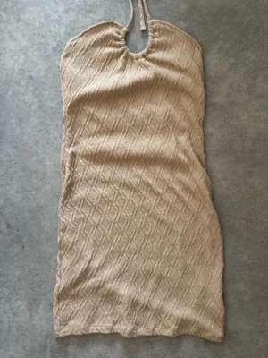Beige stickad halterneckklänning - Snygg beige kort stickad klänning med halterneck och knyt i nacken. Klänningen har ett diskret mönster i tyget och är perfekt för varma dagar. Mjuk och bekväm med en lätt figurnära passform.