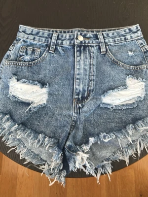 Blå jeansshorts med fransar - Säljer ett par blå jeansshorts med slitna detaljer och fransig kant. Shortsen har klassisk femficksdesign och råa hål framtill för en edgy look. Perfekta till sommaren och ger en avslappnad vibe. Helt nya 