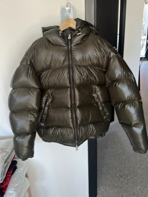 Olivgrön pufferjacka med huva - Snygg olivgrön pufferjacka med glansig finish och stor huva. Perfekt för kalla vinterdagar och ger en cool streetwear-look. Knappt använd Storlek M