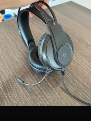 Svarta SG Gaming headset med mikrofon - Säljer ett par svarta over-ear gaminghörlurar från SG med inbyggd mikrofon och vadderade kåpor. De har en justerbar huvudbygel och ansluts med kabel. Perfekta för gaming eller videosamtal.