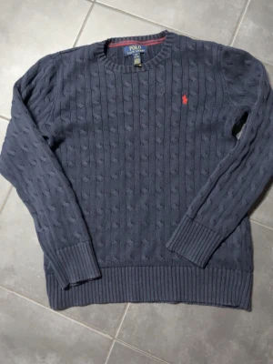 Mörkblå kabelstickad tröja Polo Ralph Lauren - Snygg mörkblå kabelstickad tröja från Polo Ralph Lauren med klassisk röd logga broderad på bröstet. Tröjan har rund hals, långa ärmar och ribbade muddar. Perfekt för dig som gillar stilrena och tidlösa plagg.