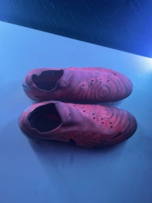 Rosa Nike fotbollsskor utan snörning - Säljer ett par rosa Nike fotbollsskor med innovativ design utan snörning och mönstrad ovandel. Skorna har rundade former och tydliga texturer för bättre bollkontroll. Sulan är ljusrosa med fasta dobbar och Nike-logga. Perfekta för spel på gräs.