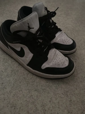 Svarta och vita Air Jordan 1 Low - Säljer ett par Air Jordan 1 Low i svart och vitt läder med klassisk Nike Swoosh på sidan och Jumpman-logga på plösen. Skorna har perforerad tåbox för ventilation och svarta skosnören. Perfekt för dig som gillar streetwear och ikoniska sneakers.