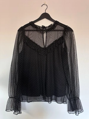 Svart transparent prickig blus - Svart blus i transparent mesh med små vita prickar och volangdetaljer vid halsen och ärmsluten. Blusen har långa ärmar och knytning i nacken. Perfekt för dig som gillar en trendig och luftig look.
