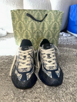 Gucci sneakers med blå GG-mönster - Snygga sneakers från Gucci med klassiskt blått GG-monogram på canvas och detaljer i mörkblått läder. Storlek: 42-43. Vita skosnören och robust sula med stjärndetaljer undertill. Märkeslogga på plösen och insidan. Perfekt för dig som vill sticka ut med exklusiv design.
