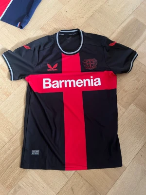 Bayer Leverkusen matchtröja svart/röd - Snygg Bayer Leverkusen matchtröja från Castore i svart med röd korsdesign och vita detaljer. Tryckt sponsorlogga 'Barmenia' på bröstet och klubbmärke. Tillverkad i lätt och ventilerande funktionsmaterial, perfekt för fotboll eller träning.