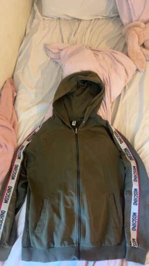 Olivgrön hoodie från Moschino - Snygg olivgrön hoodie från Moschino med dragkedja framtill och vita band med Moschino-logga längs ärmarna. Hoodien har huva, ribbade muddar och två fickor framtill. Perfekt för en avslappnad och trendig look.