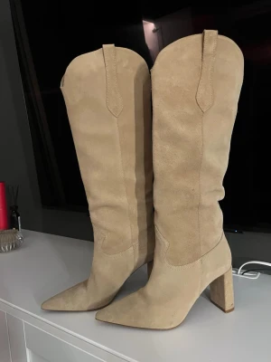 Beige höga cowboy boots i mocka - Säljer ett par snygga beige cowboy boots i mocka med spetsig tå och bred klack. Den har endast använts en gång så i bra skick. Ny pris:1259kr 