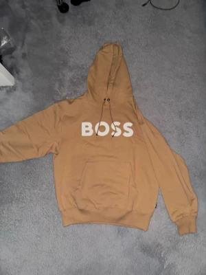 Boss hoodie  - Snygg beige hoodie från BOSS med stor vit logga på bröstet. Hoodien har dragsko i huvan och en klassisk känguruficka framtill. Perfekt för en avslappnad och trendig stil.