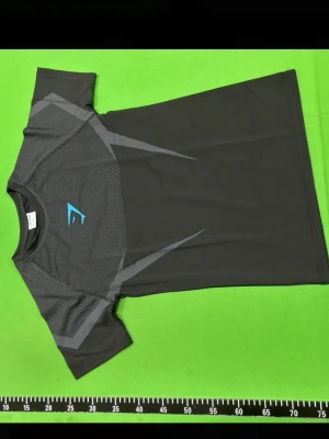 Gymshark t-shirt - Snygg svart sporttröja från gymshark med grå mönstrade detaljer och blå logga på bröstet. Tillverkad i ett lätt och snabbtorkande syntetmaterial som passar perfekt till träning. Kortärmad och med modern passform.