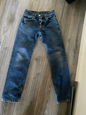Blå raka Levi's 501 jeans med knappar - Klassiska blå jeans från Levi's med rak passform och knappgylf. Jeansen har fem fickor, kopparfärgade nitar och kontrastsömmar. Tillverkade i slitstark denim som ger en tidlös look. Perfekta för dig som gillar en avslappnad och stilren stil. W27 L30