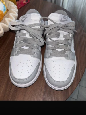 Nike Dunk Low grå/vita sneakers strl 36 - Nike Dunk Low sneakers i stilren grå och vit färg. Skorna har klassisk låg siluett, perforerad tåbox och ikoniska swoosh-loggan på sidan. Tillverkade i läder med textildetaljer och matchande grå skosnören. Perfekta för dig som gillar clean och tidlös streetstil. Använda 2 gånger, säljes pga försmå!