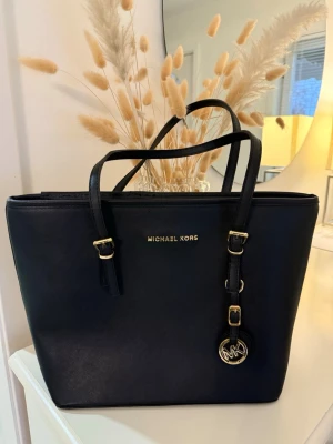 Svart handväska från Michael Kors - Inte äkta. Stilren svart handväska från Michael Kors i slätt läder med guldfärgade detaljer och logotyp framtill. Väskan har dubbla axelremmar och ett dekorativt MK-hänge. Perfekt för dig som vill ha en klassisk och trendig accessoar. 