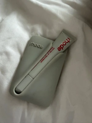Rhode Peptide Lip Treatment - Rhode Peptide Lip Treatment i en stilren tub med röd text och grått fodral. Läppbalsamet har en minimalistisk design och är perfekt för att återfukta och vårda läpparna. Tuben är gjord av plast och har en praktisk applikator.