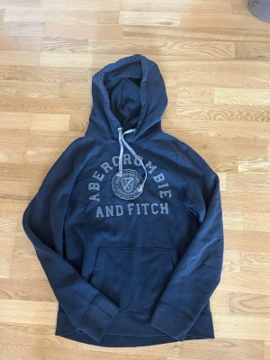 Mörkblå hoodie Abercrombie & Fitch - Mörkblå hoodie från Abercrombie & Fitch med stor logga och text framtill. Klassisk känguruficka och justerbar huva med vita snören. Skön och avslappnad passform, perfekt till vardagslooken. Tillverkad i mjukt bomullsmaterial.