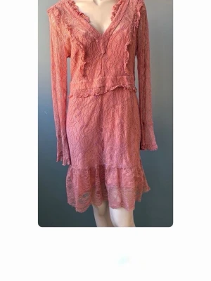 Rosa spetsklänning med volang - Romantisk rosa klänning i spets med v-ringning och volangdetaljer. Klänningen har lång ärm och är tillverkad i ett tunt, genombrutet material som ger en feminin känsla. Perfekt för dig som gillar en söt och trendig stil. Storlek XL men sitter som M. 