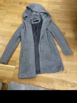 Grå lång kappa med huva - Snygg grå kappa med huva och dragkedja framtill. Kappan är fodrad och har en rak passform med långa ärmar. Perfekt för kyliga dagar och enkel att matcha med olika outfits.