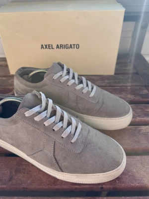 Gråa sneakers från Axel Arigato - Snygga gråa sneakers från Axel Arigato i mocka med vita snören och vit sula. Klassisk låg modell med stilren design som passar till det mesta. Perfekta för dig som gillar minimalistisk och trendig stil.