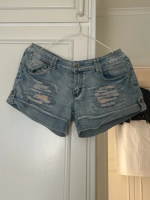 Blå slitna jeansshorts - Säljer ett par blå jeansshorts med slitna detaljer framtill och klassisk femficksdesign. Shortsen har knapp- och dragkedjestängning samt uppvikta benslut. Perfekta för sommaren och ger en avslappnad look.