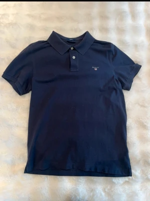 Mörkblå pikétröja från Gant - Klassisk mörkblå pikétröja från Gant med korta ärmar och krage. Tröjan har två knappar framtill och en liten broderad Gant-logga på bröstet. Perfekt för en stilren och avslappnad look.