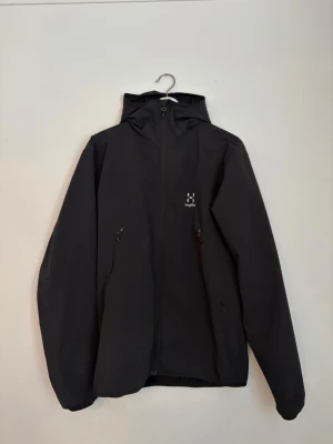 Svart vindjacka från Haglöfs - Svart vindjacka från Haglöfs med minimalistisk design, hel dragkedja framtill och två fickor med dragkedja. Jackan har huva och är tillverkad i ett lätt och slitstarkt material, perfekt för blåsiga dagar. Diskret Haglöfs-logga på bröstet.