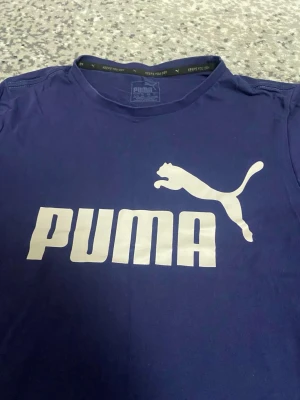Marinblå Puma t-shirt med logga - Snygg marinblå t-shirt från Puma med stor vit logga och text på bröstet. Klassisk rund hals och korta ärmar. Tillverkad i mjuk bomull som känns skön mot huden. Perfekt för dig som gillar sportig och enkel stil.