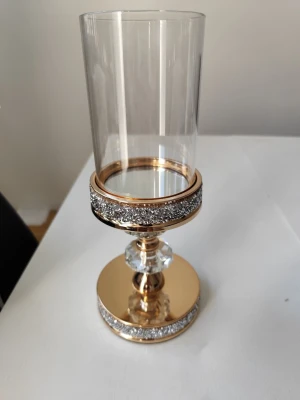 Glaslykta med guld och strassdetaljer - Snygg glaslykta med cylinderformad glascylinder, guldfärgad metallfot och dekorativa strassdetaljer. Foten har två stora klara kristalliknande dekorationer och glittrande band runt både fot och överkant. Perfekt som inredningsdetalj för dig som gillar bling.