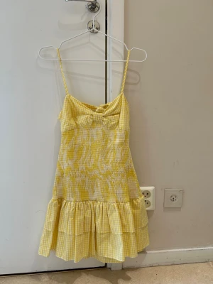  Ny slutsåld Gul rutig klänning med rosett - Söt kortklänning i gul och vit rutigt mönster med volang nedtill och rosett både fram och bak. Klänningen har smockad midja, tunna axelband och öppen rygg med knyt. Perfekt för sommarens dagar!