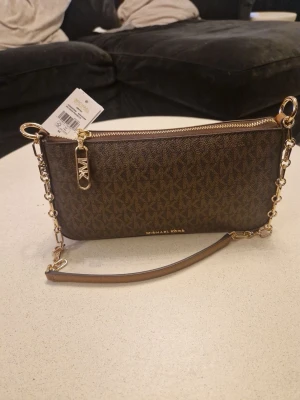 Brun Michael Kors handväska med kedja - Snygg brun handväska från Michael Kors med klassiskt MK-mönster. Väskan har guldfärgade detaljer, kedjerem och dragkedja med MK-logga. Perfekt storlek för det viktigaste och en stilren accessoar till din outfit.