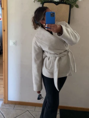 Beige omlottkappa med knytskärp från Vero Moda  - Stilren beige omlottkappa med bred krage och knytskärp i midjan. Passar perfekt på vår och höst🥰