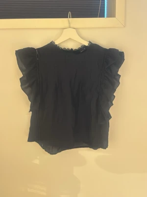 Svart volangblus med spetsdetalj - Svart blus med volanger längs ärmhålen och fin spetskant vid halsringningen. Blusen är ärmlös och har en luftig, lätt transparent känsla. Perfekt för en trendig och chic look.