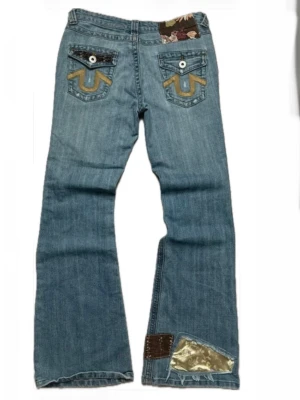 True Religion jeans med patchdetaljer - Unika ljusblå jeans från True Religion med broderade bakfickor, vita knappar och coola patchdetaljer i gult, brunt och guld. Blommigt tyg syns vid linningen och en metallic patch nertill på benet. 