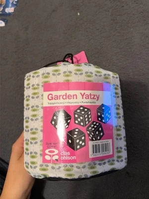 Garden Yatzy från Clas Ohlson - Trädgårdsyatzy från Clas Ohlson i en vit och grönmönstrad tygpåse med dragsko. Perfekt för utomhusspel med stora svarta tärningar och tydliga vita prickar. Kommer i en praktisk förvaringspåse med handtag.