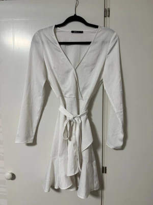 Vit omlottklänning med bälte - Snygg vit omlottklänning från Gina Tricot med lång ärm och v-ringning. Klänningen har ett knytbart bälte i midjan. Perfekt till studenten eller sommaren. Aldrig använd. 