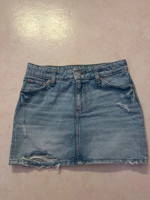 Ljusblå jeanskjol med slitningar - Trendig ljusblå jeanskjol med rå kant och slitna detaljer framtill. Klassisk femficksmodell med knapp och dragkedja. Perfekt för en avslappnad och cool stil.