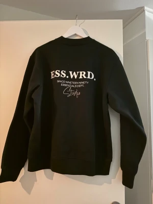 Svart sweatshirt ESS WRD Studio - Cool svart sweatshirt från ESS WRD Studio med vit text på ryggen. Tröjan har rund halsringning och långa ärmar. Perfekt för dig som gillar streetwear och enkel stil.