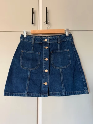 Jeanskjol med knappar från River Island - Snygg blå jeanskjol från River Island med knappar framtill och två fickor fram o bak. Kjolen har en klassisk A-linjeform och bälteshällor i midjan. 