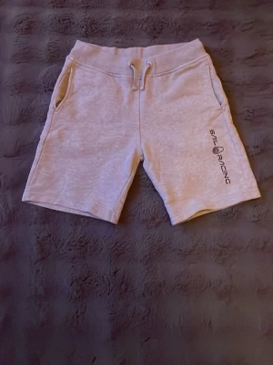Gråa shorts från Sail Racing - Snygga ljusgrå shorts från Sail Racing med dragsko i midjan, två sidofickor och en bakficka. Mjukt material som känns skönt mot huden och tryckt logga på ena benet. Perfekta för en avslappnad stil.