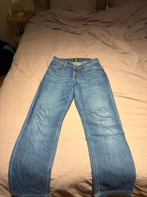 Blå raka jeans från Lee - Säljer ett par klassiska blå jeans från Lee med rak passform och fem fickor. Jeansen har en ljus tvätt och Lee-logga på bakfickan. Tillverkade i slitstarkt denimtyg, perfekta för en avslappnad och tidlös stil. Längd på jeans är 28 / 30 för hår sytt upp dom.