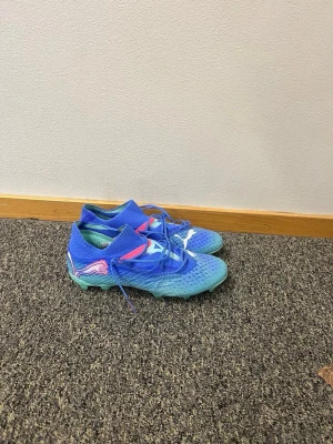 Blåa Puma fotbollsskor med mönster - Säljer ett par blåa fotbollsskor från Puma med vita och rosa detaljer samt stickad strumpliknande krage. Skorna har snörning och greppvänlig sula med dobbar, perfekt för spel på gräs. Ovandelen är i syntetmaterial med ventilerande mesh.