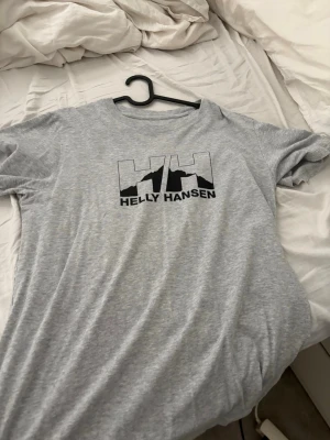 Grå Helly Hansen t-shirt med logga - Snygg grå t-shirt från Helly Hansen med stor svart logga på bröstet. Klassisk passform och rund halsringning
