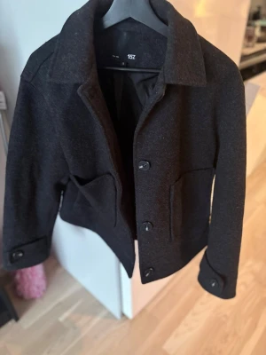 Svart kort jacka från 157 - Stilren svart jacka från 157 med klassisk krage, knäppning framtill och två stora fickor. Jackan är kortare i modellen och har knappar vid ärmsluten. Perfekt för dig som gillar en enkel och snygg look.